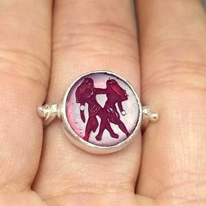 May include: A silver ring with a pink cameo featuring a carved image of two figures. The cameo is set in a round bezel.