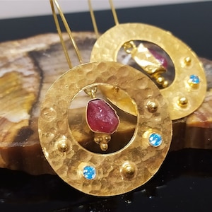 Könnte beinhalten: Goldene Ohrringe mit einem gehämmerten runden Design. Jeder Ohrring hat einen roten Edelstein und drei blaue Edelsteine.