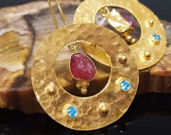 Può includere: Orecchini d'oro con un designo circolare martellato. Ogni orecchino presenta una pietra preziosa rossa e tre pietre preziose blu.