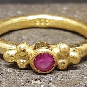 May include: A gold ring featuring a red gemstone and three small gold balls on either side of the stone.