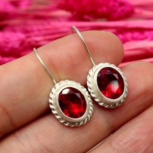 Peut inclure: Une paire de boucles d'oreilles en argent avec des pierres précieuses ovales rouges. Les boucles d'oreilles ont un design en forme de corde autour du bord du sertissage.