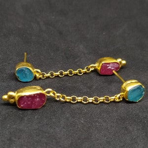 Op de afbeelding: Een paar goudkleurige oorbellen met een spijker- en kettingontwerp. Elke oorbel heeft een goudkleurige zetting met een rechthoekige, ruw geslepen edelsteen. Eén edelsteen is turquoise, de andere is diep magenta.