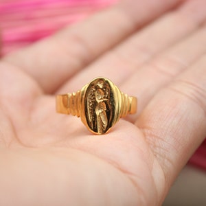 Op de afbeelding: Een gouden ring met een ovaalvormig ontwerp met een engel met vleugels. De ring is op de hand van een persoon.