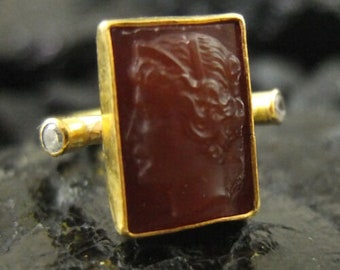 May include: A gold ring with a rectangular carnelian stone featuring a carved profile of a woman. The ring has two small diamond accents on the band.