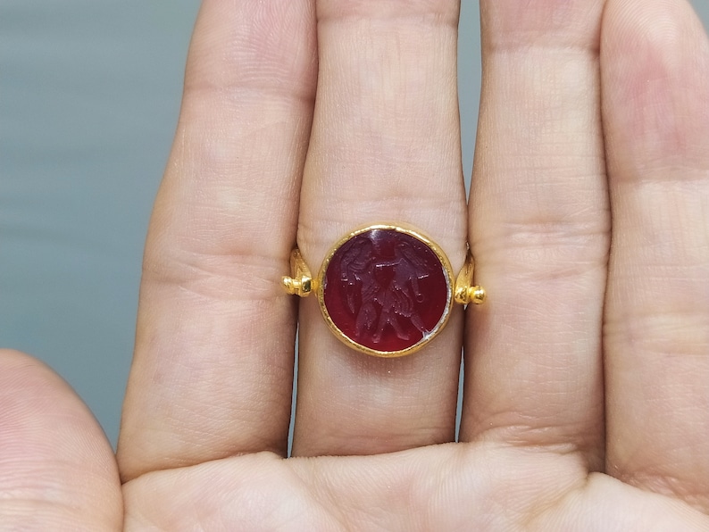 Peut inclure: Une bague en or avec une pierre de cornaline rouge orn&eacute;e d'un motif sculpt&eacute; de deux figures. La bague a une bande simple et une pierre ronde.