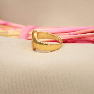 May include: A gold signet ring with a smooth, rounded band. The ring is lying on a light pink and white surface.