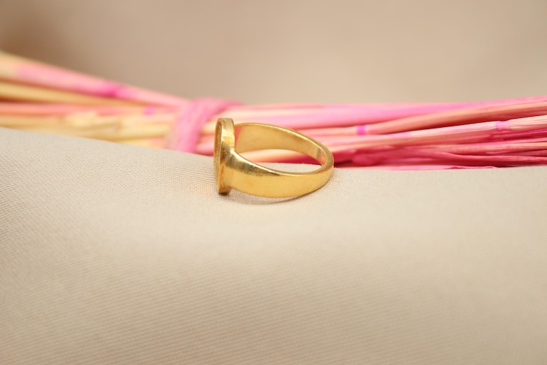 May include: A gold signet ring with a smooth, rounded band. The ring is lying on a light pink and white surface.