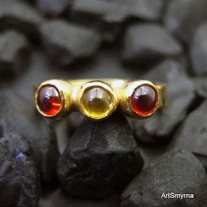 May include: A gold ring with three gemstones. The gemstones are red, yellow, and red. The ring is on a black background.