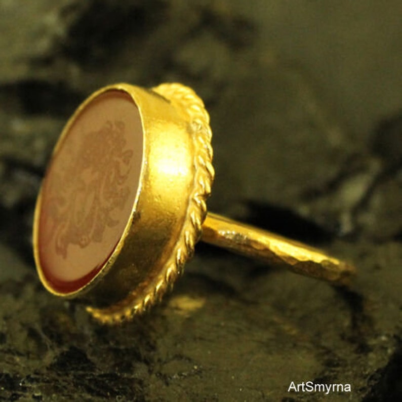 May include: A gold ring with an oval, reddish-brown stone. The stone has an engraved design. The ring has a twisted rope-like border around the stone and a hammered band. The ring is set against a dark background.