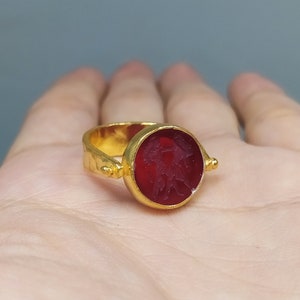 Peut inclure: Une bague en or avec une pierre pr&eacute;cieuse rouge sertie dans un sertissage &agrave; b&eacute;li&egrave;re. La pierre pr&eacute;cieuse a un motif grav&eacute;.