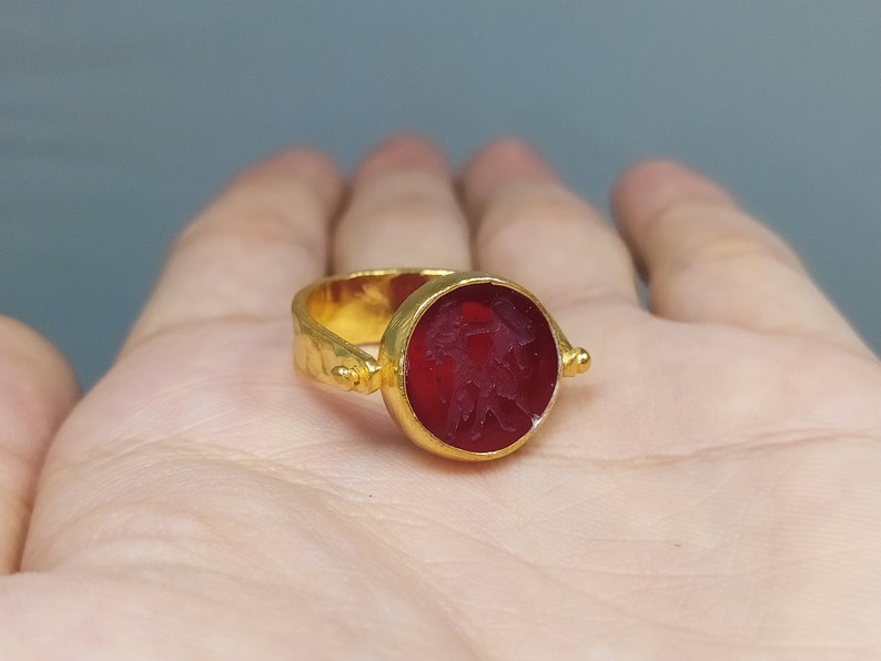 Peut inclure: Une bague en or avec une pierre pr&eacute;cieuse rouge sertie dans un sertissage &agrave; b&eacute;li&egrave;re. La pierre pr&eacute;cieuse a un motif grav&eacute;.