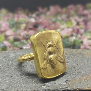 May include: A gold ring with a rectangular face featuring a detailed embossed image of a bee.