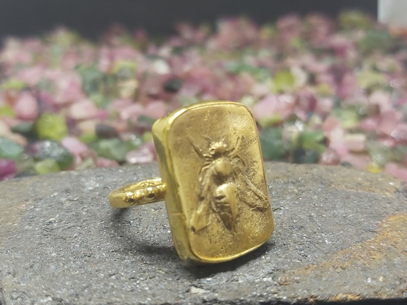 May include: A gold ring with a rectangular face featuring a detailed embossed image of a bee.