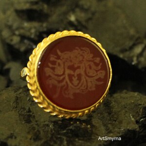 May include: A gold-colored ring with a round, dark red stone. The stone features a detailed, light-colored design in the center. The ring's band has a textured, rope-like design around the stone. The ring is set against a dark, textured background.