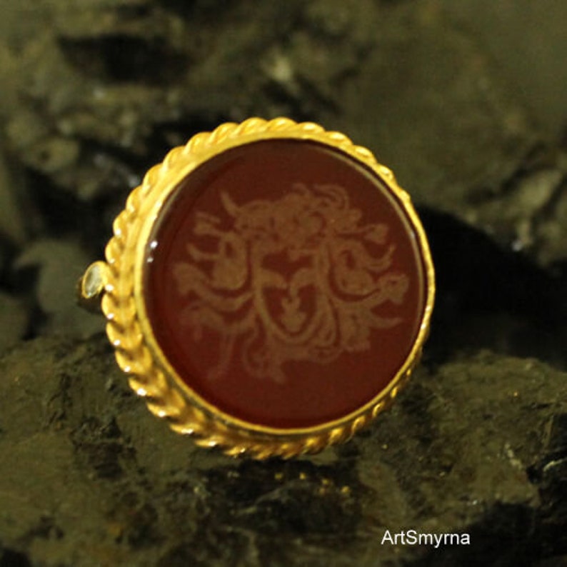 May include: A gold-colored ring with a round, dark red stone. The stone features a detailed, light-colored design in the center. The ring's band has a textured, rope-like design around the stone. The ring is set against a dark, textured background.