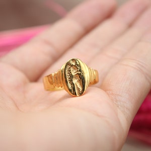 Op de afbeelding: Een gouden ring met een ovaalvormig ontwerp met een gedetailleerde gravure van een persoon. De ring is op de hand van een persoon.
