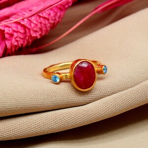 K&ouml;nnte beinhalten: Ein goldener Ring mit einem gro&szlig;en ovalen roten Edelstein und zwei kleineren blauen Edelsteinen.