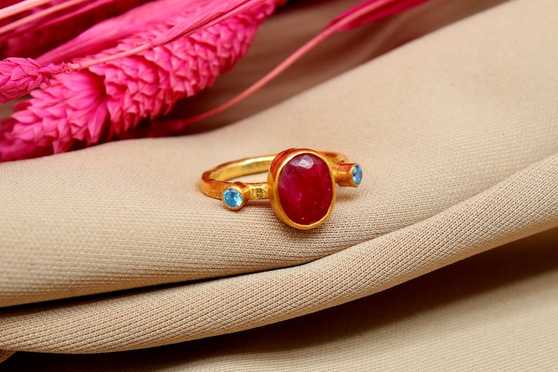 K&ouml;nnte beinhalten: Ein goldener Ring mit einem gro&szlig;en ovalen roten Edelstein und zwei kleineren blauen Edelsteinen.