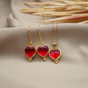 May include: A set of gold heart-shaped earrings and a necklace with red glass stones. The jewellery is on a light brown fabric background.