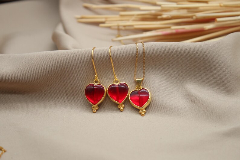 May include: A set of gold heart-shaped earrings and a necklace with red glass stones. The jewellery is on a light brown fabric background.