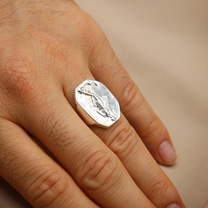 May include: A silver ring with a rectangular face featuring a floral design. The ring is worn on a finger, showcasing its polished metal finish. The background is a neutral beige.