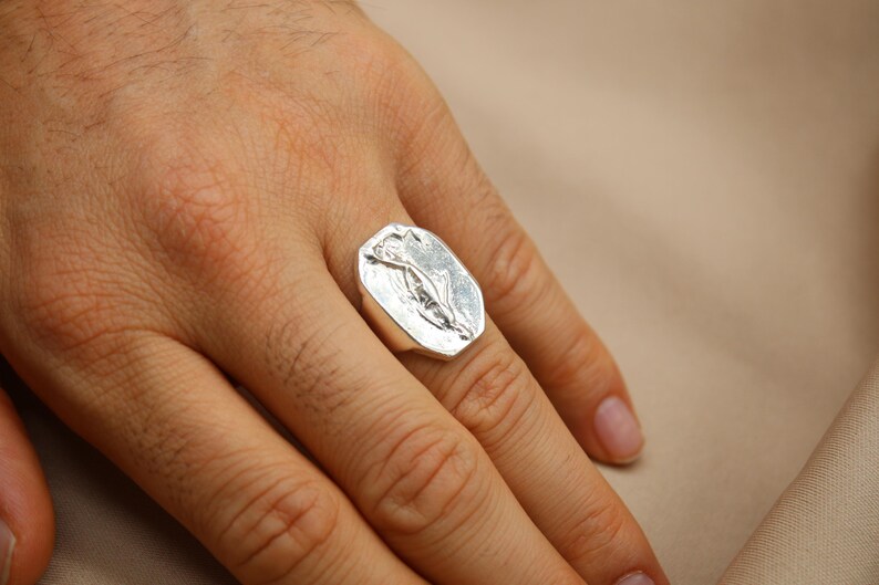 May include: A silver ring with a rectangular face featuring a floral design. The ring is worn on a finger, showcasing its polished metal finish. The background is a neutral beige.