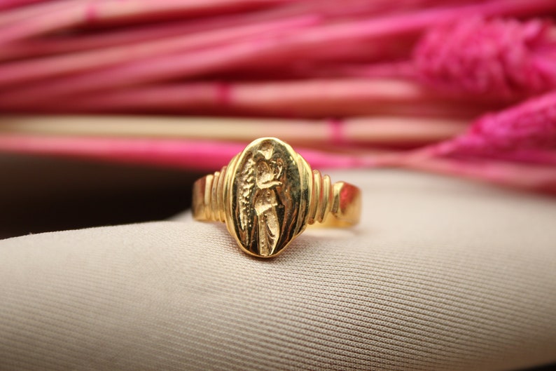 Op de afbeelding: Een gouden ring met een gedetailleerd ontwerp van een figuur met vleugels. De ring is geplaatst tegen een roze achtergrond.