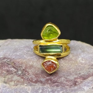 Könnte beinhalten: Goldring mit drei Edelsteinen: einem grünen Stein, einem rechteckigen grünen Stein und einem roten Stein.