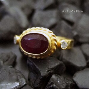 K&ouml;nnte beinhalten: Ein goldener Ring mit einem gro&szlig;en ovalen Rubin-Edelstein und zwei kleineren Diamanten. Der Ring hat eine strukturierte Band.