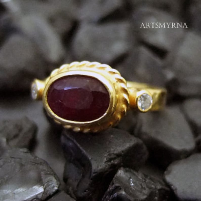 K&ouml;nnte beinhalten: Ein goldener Ring mit einem gro&szlig;en ovalen Rubin-Edelstein und zwei kleineren Diamanten. Der Ring hat eine strukturierte Band.
