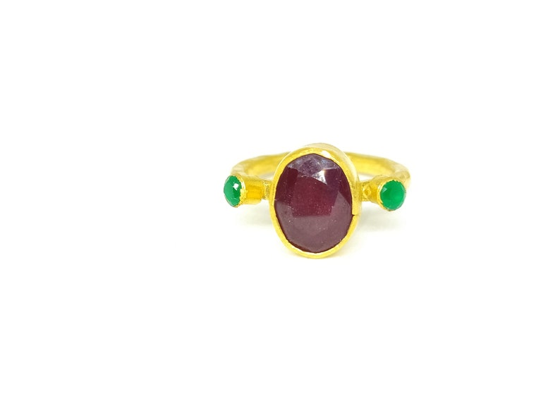 May include: A gold ring featuring a large oval ruby gemstone, flanked by two smaller green gemstones.