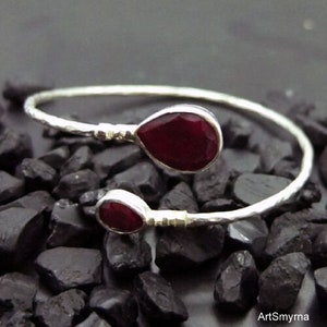 May include: A silver bangle bracelet with two teardrop-shaped red gemstones. The bracelet has a hammered texture.