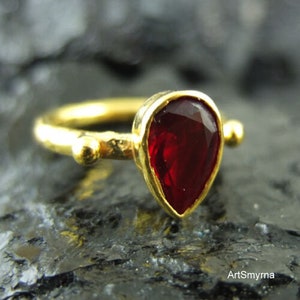 May include: A gold ring with a teardrop-shaped red gemstone. The ring has a hammered texture and two small gold balls on either side of the stone.