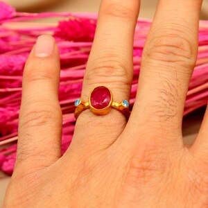 K&ouml;nnte beinhalten: Ein goldener Ring mit einem gro&szlig;en ovalen roten Edelstein und zwei kleineren blauen Edelsteinen an beiden Seiten.