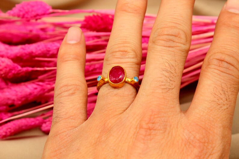 K&ouml;nnte beinhalten: Ein goldener Ring mit einem gro&szlig;en ovalen roten Edelstein und zwei kleineren blauen Edelsteinen an beiden Seiten.