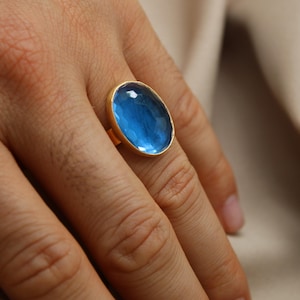 May include: A gold ring with a large oval blue gemstone. The ring is on a person's finger.