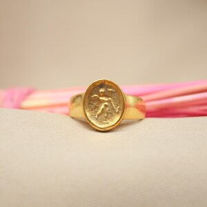 May include: A gold ring with an oval-shaped design featuring a raised image of a person running. The ring is on a pink and white background.