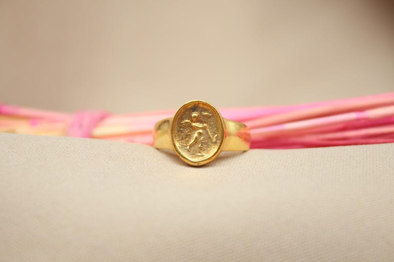 May include: A gold ring with an oval-shaped design featuring a raised image of a person running. The ring is on a pink and white background.