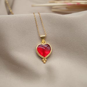 Puede incluir: Un collar de cadena dorada con un colgante de corazón rojo. El corazón está engastado en oro y tiene un diseño detallado.