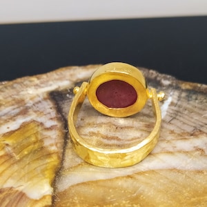 Peut inclure: Une bague en or avec une pierre ovale rouge fonc&eacute; sertie dans un sertissage &agrave; b&eacute;li&egrave;re. La bande de la bague est textur&eacute;e et a une finition martel&eacute;e.