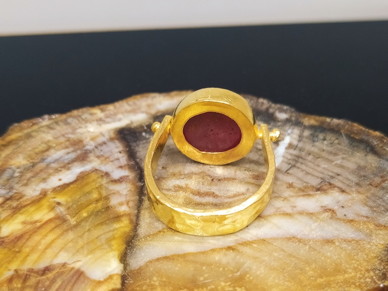 Peut inclure: Une bague en or avec une pierre ovale rouge fonc&eacute; sertie dans un sertissage &agrave; b&eacute;li&egrave;re. La bande de la bague est textur&eacute;e et a une finition martel&eacute;e.
