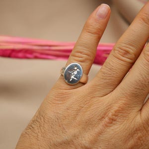 以下が含まれることがあります： 花柄の楕円形のデザインが施されたシルバーリング。リングは手の指先につけられています。