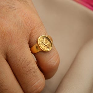 May include: A gold ring with a round, raised design featuring a stylized bird. The ring is worn on a person's finger.