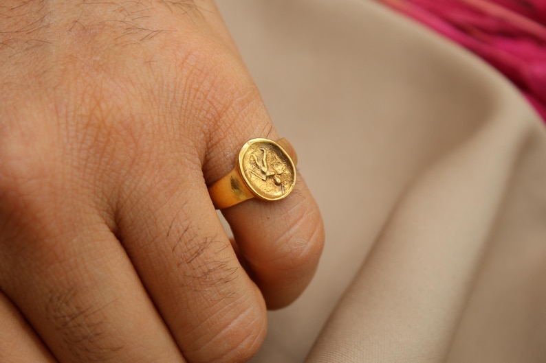 May include: A gold ring with a round, raised design featuring a stylized bird. The ring is worn on a person's finger.