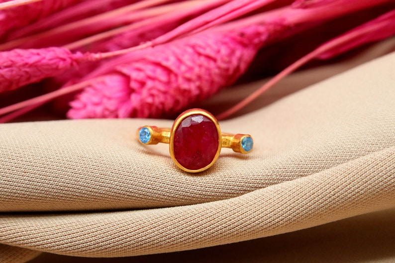 K&ouml;nnte beinhalten: Ein goldener Ring mit einem gro&szlig;en ovalen roten Edelstein und zwei kleineren blauen Edelsteinen an beiden Seiten.