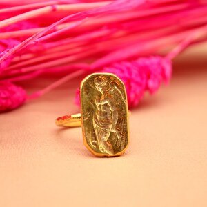May include: A gold ring with a rectangular design featuring an embossed image of an angel with wings.