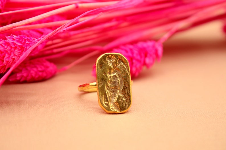 May include: A gold ring with a rectangular design featuring an embossed image of an angel with wings.