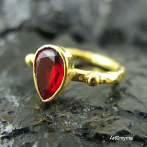 May include: A gold ring with a teardrop-shaped red gemstone. The ring band is textured and has small gold beads.
