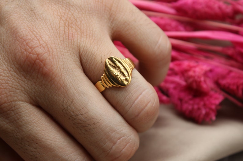 Op de afbeelding: Een gouden ring met een getextureerd ovaalvormig ontwerp. De ring wordt aan een vinger gedragen.