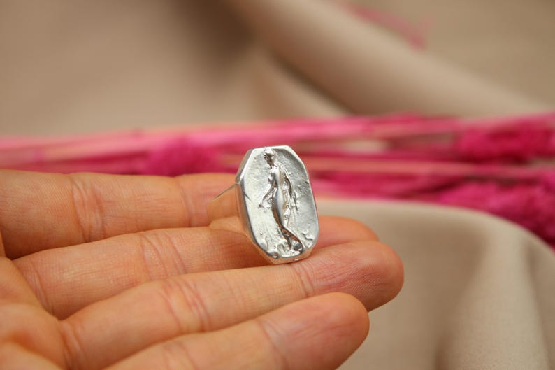 May include: A silver ring with a rectangular face featuring a sculpted image of a woman. The ring is held in a hand, with a blurred background of pink and beige. The ring's design is detailed and the metal has a slightly textured appearance.